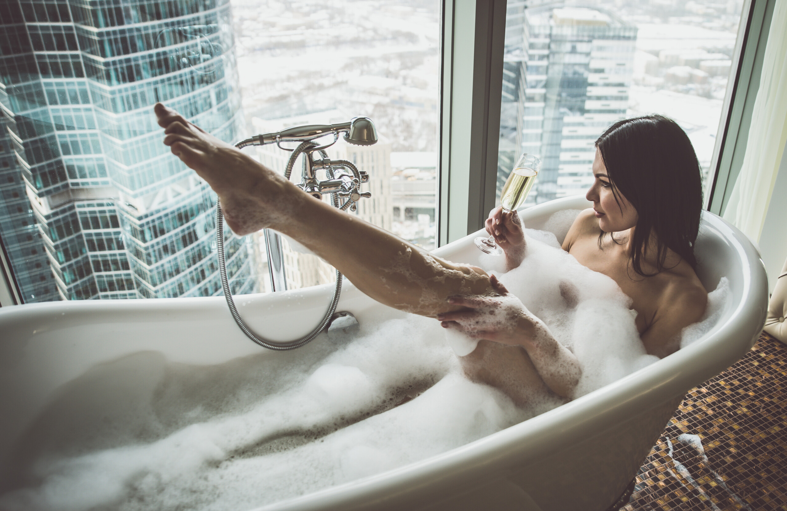 dark haired woman drinks champagne in the bath looking out at the london city skyline